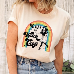 Pode incluir: Uma camiseta creme com o texto "BE GAY DO Witch Craft" acima de um arco-&iacute;ris e um gr&aacute;fico de bruxa. A bruxa est&aacute; usando um chap&eacute;u e uma saia. A pessoa est&aacute; usando jeans azuis.