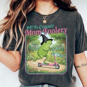 Puede incluir: Camiseta gris oscuro con el texto "Off To Commit Mom Foolery". La camiseta presenta una rana de dibujos animados con un sombrero de bruja y montada en un patinete rosa. La rana está en un parque con árboles y un estanque.