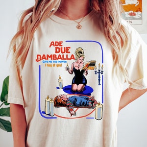 Może przedstawiać: Kremowa koszulka z nadrukiem z napisem "ADE DUE DAMBALLA GIVE ME THE POWER I beg of you!". Grafika przedstawia kobietę w czarnej sukience, świece i lalkę. Jest to element odzieży codziennej.