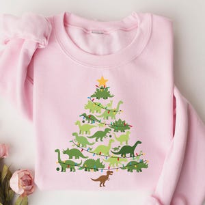 Puede incluir: Sudadera rosa claro con un diseño de árbol de Navidad hecho de dinosaurios verdes. El árbol está coronado con una estrella amarilla y decorado con luces de cadena. Un dinosaurio marrón está en la base del árbol.