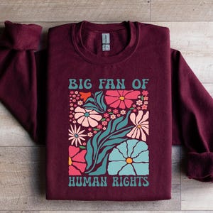 May include: Burgundy sweatshirt with the text "BIG FAN OF HUMAN RIGHTS" in teal and white. The design features a floral arrangement with pink, teal, and white flowers. The sweatshirt is made of a soft material.