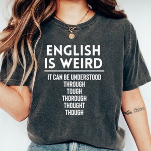 May include: A dark gray t-shirt with the text "ENGLISH IS WEIRD" and the words "IT CAN BE UNDERSTOOD THROUGH TOUGH THOROUGH THOUGHT THOUGH" printed in white.