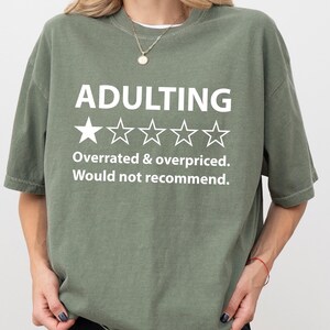 May include: A green t-shirt with the text "Adulting" and a one-star rating. The text below the rating reads "Overrated & overpriced. Would not recommend."
