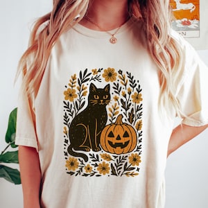 May include: Cream-colored t-shirt featuring a black cat and jack-o'-lantern design. The graphic is surrounded by floral and leaf elements in shades of gold and brown. A Halloween-themed tee.