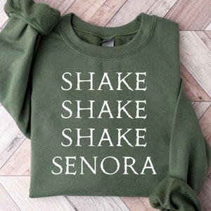 May include: An olive green sweatshirt with the words "SHAKE SHAKE SHAKE SENORA" printed in white. The sweatshirt has a crew neck and long sleeves with ribbed cuffs. The design is centered on the front of the garment.