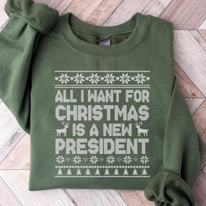 以下が含まれることがあります： オリーブグリーンのスウェットシャツ。白で「ALL I WANT FOR CHRISTMAS IS A NEW PRESIDENT」と書かれています。雪の結晶とトナカイのモチーフがデザインされ、お祝いのニットセーターのようです。