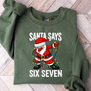 May include: An olive green sweatshirt with the text "SANTA SAYS SIX SEVEN" in white. The graphic features Santa Claus dabbing, wearing a red suit and hat, and wrapped in Christmas lights.