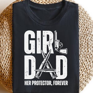 May include: Black t-shirt with white text and graphics. The text reads "GIRL DAD HER PROTECTOR, FOREVER." The design includes a handgun and crossed bullets. The shirt is displayed on a woven, circular surface.