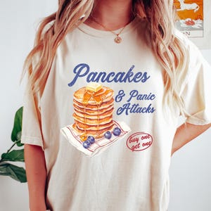 May include: A cream-coloured t-shirt with the text "Pancakes & Panic Attacks" in blue script. The graphic shows a stack of pancakes with butter, blueberries, and the phrase "buy one get one".