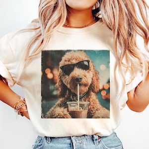 May include: Cream-colored t-shirt featuring a graphic of a dog wearing sunglasses and drinking from a coffee cup with a straw. The dog is a light brown color. The background is blurred with bokeh lights.