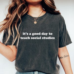 Op de afbeelding: Donkergrijs t-shirt met de witte tekst "it's a good day to teach social studies". Het shirt wordt gecombineerd met een blauwe jeans en witte sneakers. Het thema is onderwijs en lesgeven.