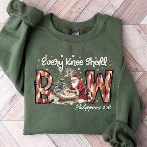 May include: An olive green sweatshirt with the text "Every Knee Shall Bow" in gold lettering. The letters "B" and "W" are in a camouflage pattern. A graphic of Santa Claus and baby Jesus is also featured.