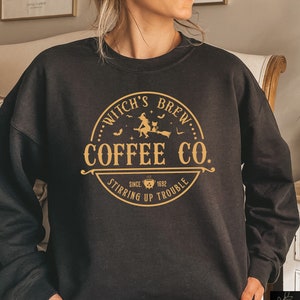 以下が含まれることがあります： 黒いクルーネックのスウェットシャツ。金色のグラフィックで「Witch's Brew Coffee Co. Stirring Up Trouble Since 1692」と書かれており、ほうきに乗った魔女、コウモリ、猫が描かれています。快適でカジュアルなスタイルです。