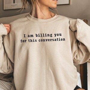 Może przedstawiać: Beżowa bluza z czarnym napisem "I am billing you for this conversation". Top z długim rękawem jest w neutralnym kolorze. Zdjęcie zrobiono w pomieszczeniu.