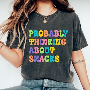Pode incluir: T-shirt cinza escuro com o texto "PROBABLY THINKING ABOUT SNACKS" em letras coloridas. A camisa é de estilo casual, adequada para o uso diário. A modelo está usando jeans azuis e tênis brancos.