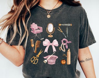 May include: Dark gray t-shirt featuring watercolor illustrations of salon tools, including a pink chair, mirror, scissors, and hair dryer. Other elements include a bow, hairbrush, and hair products. The shirt is a comfortable, casual style.