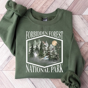 Könnte beinhalten: Ein olivgrünes Sweatshirt mit dem weißen Schriftzug "Forbidden Forest National Park". Die Grafik zeigt eine Waldszene mit Bäumen, einem Vollmond und einer nebligen Atmosphäre. Das Sweatshirt hat einen Rundhalsausschnitt und lange Ärmel.
