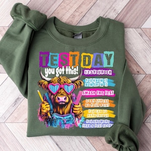 May include: An olive green sweatshirt with the text "TEST DAY you got this!" and a cartoon cow wearing heart-shaped sunglasses. The design includes colorful text blocks with motivational phrases.
