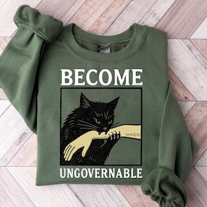 Puede incluir: Una sudadera verde oliva con las palabras "BECOME UNGOVERNABLE" impresas en blanco. El gráfico presenta un gato negro mordiendo una mano. La sudadera está hecha de un material suave.