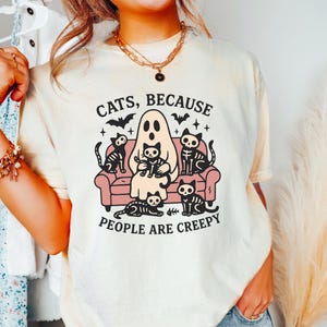 May include: Cream-coloured t-shirt with a Halloween-themed graphic. The design features a ghost, skeleton cats, and the text "CATS, BECAUSE PEOPLE ARE CREEPY". The cats are sitting on a pink sofa.