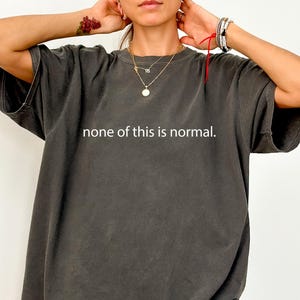 May include: Dark grey oversized t-shirt featuring the white text "none of this is normal". The shirt has short sleeves and a loose fit, suitable for casual wear.