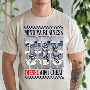 May include: Beige t-shirt featuring a graphic with three raccoons on a baseball field. The text "MIND YA BUSINESS" is above the raccoons, and "DIESEL AINT CHEAP" is below. The raccoons are in various poses.