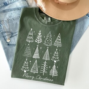 May include: An olive green t-shirt featuring a white graphic of Christmas trees and the text "Merry Christmas." The shirt is styled with light blue jeans, a gold necklace, and a tan hat with a brown band.