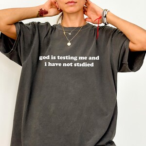 May include: Dark gray t-shirt with the text "god is testing me and i have not studied" in white. The shirt is a loose fit and has short sleeves. The shirt is a solid color.