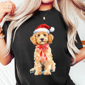 May include: Black t-shirt featuring a watercolor illustration of a golden doodle wearing a Santa hat and a red bow. The dog is sitting, with a sweet expression. The shirt has a crew neck and short sleeves. The brand name "Favour Art Co" is at the bottom.