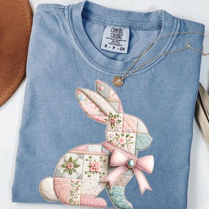 May include: A light blue Comfort Colors t-shirt featuring a patchwork bunny design with pink, white, and blue floral patterns. The bunny has a pink bow with a turquoise gem. A gold necklace with a pendant is draped on the shirt.