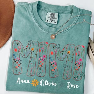 Pode incluir: Uma camiseta verde-&aacute;gua com a palavra "MAMA" em um design floral. Os nomes "Anna", "Olivia" e "Rose" s&atilde;o impressos abaixo. Um colar de prata com um pingente est&aacute; na camiseta.