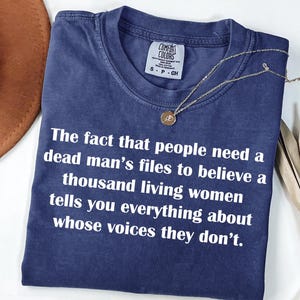 May include: A navy blue Comfort Colors t-shirt with white text: "The fact that people need a dead man's files to believe a thousand living women tells you everything about whose voices they don't." A gold necklace is draped across the shirt.