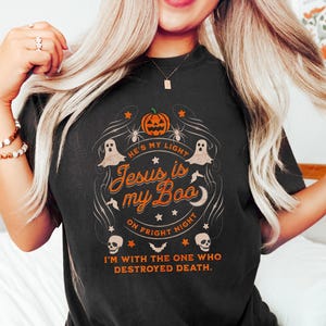 May include: Black t-shirt with a Halloween-themed graphic. The design includes a pumpkin, ghosts, spiders, and the text "Jesus is my Boo" and "On Fright Night". The shirt also has the text "I'm with the one who destroyed death."