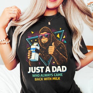 May include: Black t-shirt featuring a cartoon Bigfoot wearing sunglasses and holding a milk carton, with the text "JUST A DAD WHO ALWAYS CAME BACK WITH MILK". The design includes a retro geometric background.