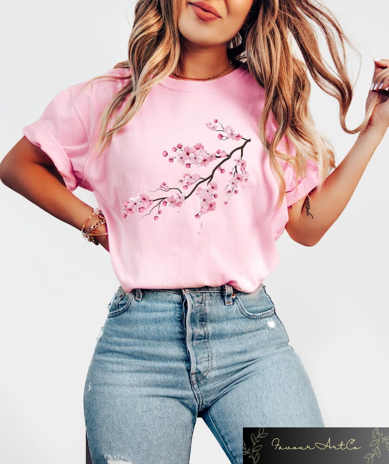 Puede incluir: Una camiseta rosa claro con una rama de flores de cerezo rosas. El gr&aacute;fico est&aacute; en la parte delantera de la camiseta. La modelo lleva vaqueros azules. La marca "Favour ArtCo" es visible en la esquina inferior derecha.