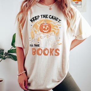 Op de afbeelding: Een wit T-shirt met een afbeelding van een pompoenkop die een boek vasthoudt. De tekst op het shirt luidt "Keep the candy, I'll take books".