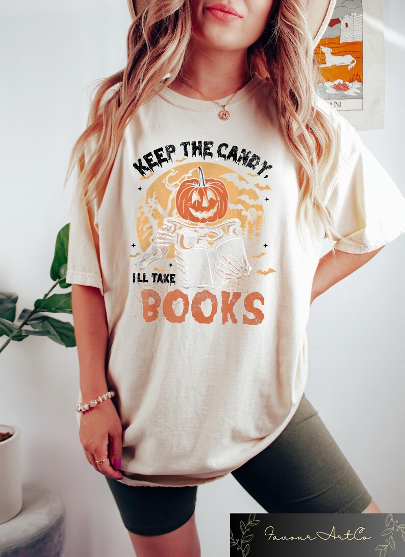 Op de afbeelding: Een wit T-shirt met een afbeelding van een pompoenkop die een boek vasthoudt. De tekst op het shirt luidt "Keep the candy, I'll take books".