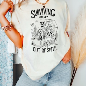 May include: A white t-shirt with a black and white graphic of a skeleton in a coffin with the text "SURVIVING PURELY OUT OF SPITE."