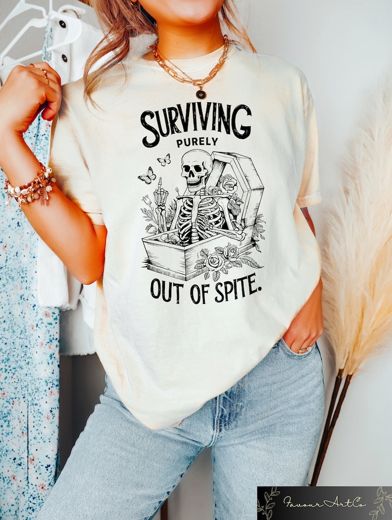 May include: A white t-shirt with a black and white graphic of a skeleton in a coffin with the text "SURVIVING PURELY OUT OF SPITE."