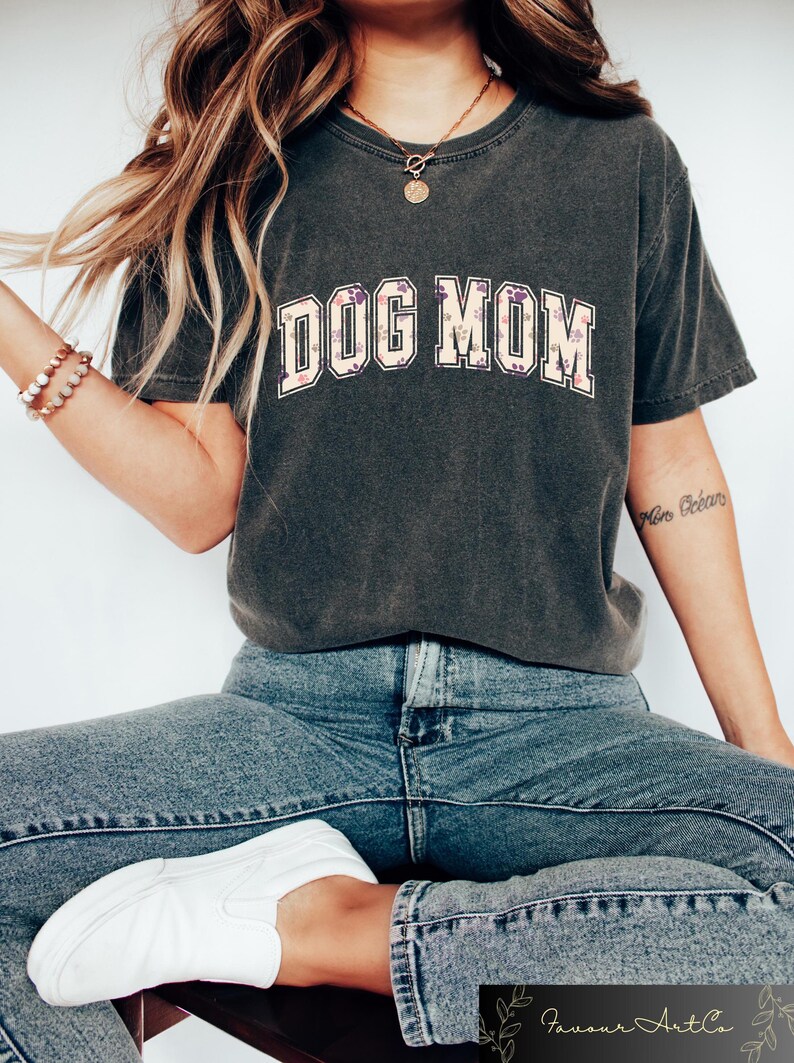 May include: A dark gray t-shirt with the text "DOG MOM" in white letters with a light beige outline. The letters are decorated with a floral pattern. The shirt is worn by a person wearing blue jeans and white sneakers.