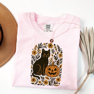 May include: A light pink Comfort Colors t-shirt featuring a black cat and jack-o'-lantern design within a floral arch. The cat has yellow eyes and sits next to the orange pumpkin. A gold necklace is draped on the shirt.