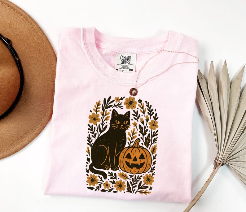 May include: A light pink Comfort Colors t-shirt featuring a black cat and jack-o'-lantern design within a floral arch. The cat has yellow eyes and sits next to the orange pumpkin. A gold necklace is draped on the shirt.