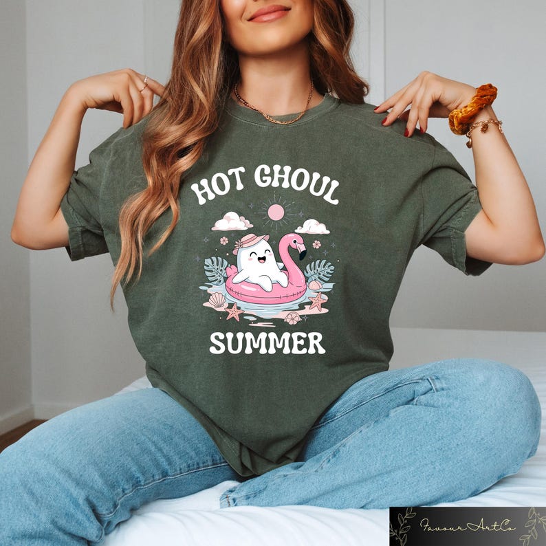 May include: Dark green t-shirt with the text "HOT GHOUL SUMMER". The graphic features a cartoon ghost in a flamingo floatie, with a sun, clouds, and ocean elements. The shirt is a relaxed fit.
