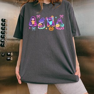 May include: A dark gray t-shirt with a neon graphic of five ghosts wearing costumes and holding candy. The ghosts are outlined in pink, blue, and green. The graphic includes a pumpkin and bows.
