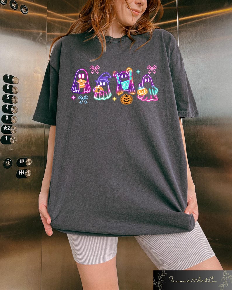 May include: A dark gray t-shirt with a neon graphic of five ghosts wearing costumes and holding candy. The ghosts are outlined in pink, blue, and green. The graphic includes a pumpkin and bows.