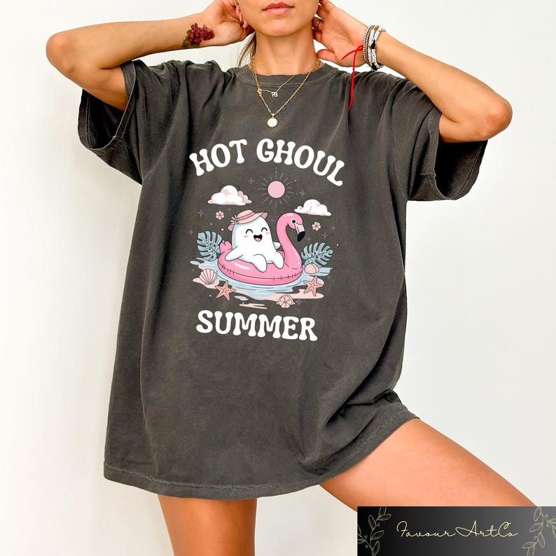 May include: Dark gray t-shirt with the words "HOT GHOUL SUMMER" in white. The graphic features a cartoon ghost in a pink flamingo floatie, with a sun, clouds, and tropical elements. The shirt is a relaxed fit.