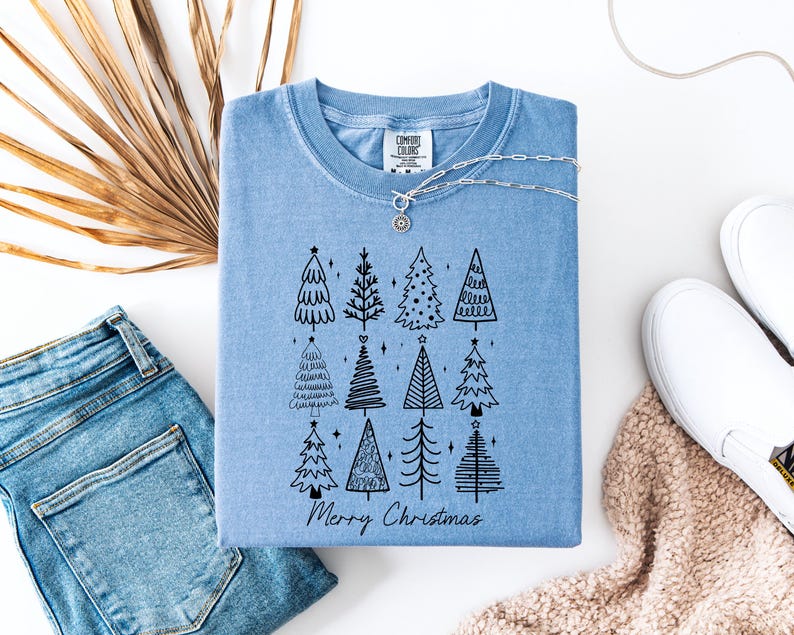 May include: A pale blue t-shirt featuring a black illustration of Christmas trees and the text "Merry Christmas." The image includes blue jeans, white trainers, and a silver necklace. A dried palm leaf is also visible.