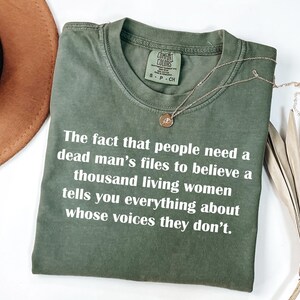 May include: Olive green t-shirt with white text: "The fact that people need a dead man's files to believe a thousand living women tells you everything about whose voices they don't." A gold necklace rests on the shirt.