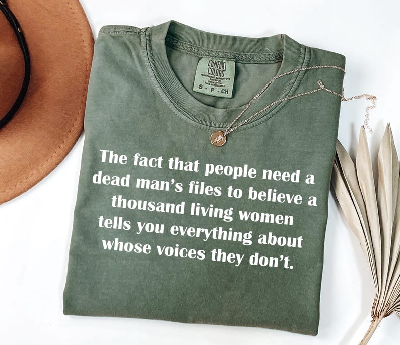 May include: Olive green t-shirt with white text: "The fact that people need a dead man's files to believe a thousand living women tells you everything about whose voices they don't." A gold necklace rests on the shirt.