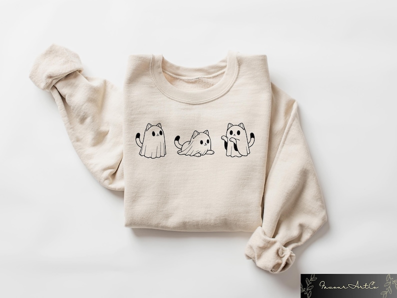 Pu&ograve; includere: Una felpa beige con una grafica nera di tre gatti vestiti da fantasmi. I gatti indossano lenzuola bianche con le code che sporgono.